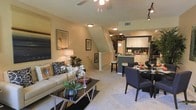 This three-story residence is designed with an open-concept layout that seamlessly connects the living, dining, and kitchen areas on the main floor. The living room is carpeted and comfortably scaled, featuring a ceiling fan, warm neutral wall tones, and thoughtfully arranged furnishings including a sofa, coffee table, and wall-mounted television. Natural and ambient lighting create a welcoming atmosphere throughout the space.

Adjacent to the living area, the dining space accommodates a full table and chairs, flowing naturally into the kitchen. The kitchen is outfitted with white cabinetry, a dark-toned island with bar seating, and modern appliances, offering both functionality and visual contrast. Track lighting above the kitchen adds a contemporary touch.

A staircase with white balusters is centrally visible, providing access to the upper and lower floors and serving as a visual anchor within the open layout. The overall design balances practicality with a warm, inviting ambiance, making the main level well-suited for both everyday living and casual entertaining.