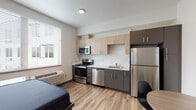 This contemporary studio unit at 3230 Kinsrow Ave in Eugene, Oregon, offers a thoughtfully designed single-floor layout that efficiently combines living, sleeping, and cooking spaces. The open-concept main area features warm wood-look flooring that flows throughout, creating a cohesive and inviting atmosphere.

The kitchen is a standout feature, outfitted with a full suite of stainless steel appliances including a range, microwave, dishwasher, and full-size refrigerator. Two-toned cabinetry in light natural wood and charcoal gray tones provides ample storage, complemented by a white tile backsplash and clean white countertops. A window beside the kitchen brings in natural light, enhancing the bright, airy feel of the space.

The sleeping area accommodates a full-size bed alongside a functional desk and chair, making it well-suited for both rest and work. Clean white walls and recessed lighting contribute to a calm, modern ambiance.

The full bathroom features a tub/shower combination with a curved rod, a single-sink vanity with a white countertop and dark wood-toned cabinetry, and wood-look flooring that ties into the rest of the unit.

A dedicated in-unit laundry closet houses a stacked washer and dryer, adding everyday convenience. A small dining area with a round table and chairs rounds out this efficiently designed, modern living space.