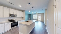 Located at 4600 Bakers Blessing Ln in Summerville, SC, this well-appointed one-bedroom, one-bathroom residence offers a thoughtfully designed single-level layout. The open-concept kitchen and living area serves as the heart of the home, featuring rich hardwood-style flooring, a large center island with a built-in sink and pendant lighting overhead, crisp white shaker cabinetry extending to the ceiling, light quartz countertops, and a full suite of stainless steel appliances. The living room receives generous natural light through large windows and a glass door, complemented by a ceiling fan overhead and a neutral color palette throughout. The bedroom is carpeted for comfort, benefits from a ceiling fan and a window with outdoor views, and connects to a spacious walk-in closet outfitted with multi-tier wire shelving. The bathroom includes a glass-enclosed walk-in shower with white tile surround and wood-look flooring. A dedicated laundry room accommodates a full-size washer and dryer and includes additional shelving for storage. Clean lines, neutral tones, and modern finishes create a bright, cohesive ambiance throughout this Summerville residence.