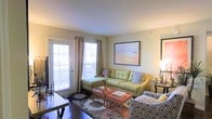This single-floor living space centers around a well-appointed living room that balances comfort and style. The room features warm, neutral-toned walls complemented by rich hardwood flooring, creating a cohesive and inviting atmosphere throughout. A large glass-paned door flanked by curtained windows allows generous natural light to fill the space, offering views to an outdoor area beyond.

The living room is furnished with a tufted chartreuse sofa paired with a matching chaise, accented by patterned throw pillows that add visual interest. A coral-toned coffee table serves as a functional centerpiece, positioned over a layered area rug that anchors the seating arrangement. Additional seating in the form of a patterned accent chair adds both comfort and character to the room.

Wall décor includes a large framed abstract artwork in warm pastel tones, enhancing the room's color palette. Ambient lighting is provided by a table lamp, supplementing the natural light. The overall ambiance is warm, casual, and thoughtfully arranged, making the space feel both functional and aesthetically pleasing.