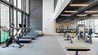 This contemporary two-floor fitness facility is designed to support a comprehensive range of training modalities within a thoughtfully appointed environment. The lower level houses the primary weight room, characterized by soaring double-height ceilings, exposed black industrial ceiling elements, and floor-to-ceiling windows that draw in natural light and offer views of the surrounding urban streetscape. This area is equipped with cable machines, power racks, weight benches, a full dumbbell rack, and stationary cycling bikes, all set on durable rubber flooring. A distinctive woven wood accent wall adds warmth and texture against the otherwise sleek, industrial aesthetic. Adjacent to the weight room is a corridor featuring the same decorative wall treatment, with seating benches, coat hooks, and a comfortable lounge nook with a sectional sofa — ideal for recovery or waiting areas. The upper level offers a bright, open functional training studio with light hardwood flooring, floor-to-ceiling glazed walls overlooking neighboring brick buildings, and an artificial turf lane outfitted with agility equipment and sled training gear. A dedicated Pilates studio with a reformer machine and corner window views completes the upper floor. A reception area with a built-in desk, restroom access, and glass entry doors provides a polished welcome point for the facility.