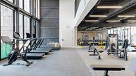 This contemporary two-floor fitness facility is designed to support a comprehensive range of training modalities within a thoughtfully appointed environment. The lower level houses the primary weight room, characterized by soaring double-height ceilings, exposed black industrial ceiling elements, and floor-to-ceiling windows that draw in natural light and offer views of the surrounding urban streetscape. This area is equipped with cable machines, power racks, weight benches, a full dumbbell rack, and stationary cycling bikes, all set on durable rubber flooring. A distinctive woven wood accent wall adds warmth and texture against the otherwise sleek, industrial aesthetic. Adjacent to the weight room is a corridor featuring the same decorative wall treatment, with seating benches, coat hooks, and a comfortable lounge nook with a sectional sofa — ideal for recovery or waiting areas. The upper level offers a bright, open functional training studio with light hardwood flooring, floor-to-ceiling glazed walls overlooking neighboring brick buildings, and an artificial turf lane outfitted with agility equipment and sled training gear. A dedicated Pilates studio with a reformer machine and corner window views completes the upper floor. A reception area with a built-in desk, restroom access, and glass entry doors provides a polished welcome point for the facility.