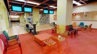 This single-floor entertainment venue is designed to deliver a lively and visually engaging experience. The bowling area features smooth, well-maintained lanes equipped with modern scoring consoles and ball return systems, each station accompanied by overhead flat-screen monitors displaying game information. The space is defined by a bold, contemporary aesthetic, with red-carpeted lounge areas furnished with translucent acrylic chairs in vivid shades of orange, yellow, and green, paired with low ottomans and round tables that create inviting social zones between lanes. A striking mixed-media wall sculpture serves as a focal point, adding an artistic dimension to the environment. The ceiling is finished with a dark grid-panel system supported by square columns, contrasted against warm yellow-toned walls that keep the atmosphere bright and energetic. Recessed and panel lighting throughout ensures excellent visibility across the facility. The open-plan layout allows for clear sightlines across the entire space, fostering a social and interactive atmosphere well-suited for group entertainment, casual outings, and recreational play.