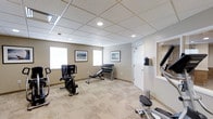 This single-level fitness room offers a practical and well-organized space designed to support a variety of workout routines. The room features a dropped ceiling with a grid tile system and recessed lighting that provides consistent, even illumination throughout. The flooring is covered with a patterned, light-toned carpet that adds a degree of comfort underfoot while maintaining a clean aesthetic.

The space is equipped with a stationary exercise bike, an elliptical machine, a weight bench, and a dumbbell rack stocked with a range of free weights, offering users options for both cardiovascular and strength training. The walls are painted in a soft, neutral beige-gray tone and are accented with framed artwork, contributing to a calm and welcoming atmosphere.

Natural light enters through multiple windows positioned along one wall, brightening the space and reducing the reliance on artificial lighting during daytime hours. A wall-mounted mirror near the entrance further enhances the sense of openness. A white panel door provides access to an adjoining area, and a small waste bin is conveniently positioned nearby. Overall, the room is clean, functional, and thoughtfully arranged.