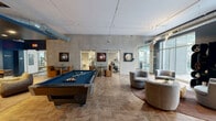 Located at 1 Franklin Town Boulevard in Philadelphia's vibrant Logan Square neighborhood, this thoughtfully designed communal living room blends industrial-chic aesthetics with sophisticated comfort. The open-plan space features striking herringbone-patterned hardwood flooring that extends throughout, complemented by bold deep-blue accent walls that lend the room a refined, contemporary character.

Exposed ductwork and track lighting run along the elevated ceilings, reinforcing the space's industrial undertones while providing ample illumination. Floor-to-ceiling glass windows and sliding doors flood the interior with natural light and offer direct views of a landscaped outdoor courtyard, seamlessly connecting the indoors to the outside environment.

The living area is furnished with curved, contemporary lounge chairs arranged around a central coffee table, creating an inviting gathering space. A tufted velvet sofa anchors one wall, beneath a curated gallery arrangement that includes a dramatic gold sunburst mirror and assorted framed artwork. A wall-mounted flat-screen television and a shuffleboard table serve as focal recreational features, while a pool table anchors the center of the room. Additional seating nooks with bold yellow accent chairs and artwork-adorned walls provide intimate corners throughout. Staircase and elevator access are also visible, ensuring excellent connectivity within the building.