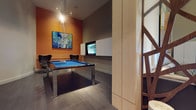 This single-floor space is thoughtfully designed as a dedicated entertainment and recreation area. The room's most prominent feature is a sleek, modern pool table with a vibrant blue felt surface and a contemporary chrome and dark wood frame, positioned centrally to allow comfortable play from all sides. Dark porcelain tile flooring grounds the space and contrasts effectively with the bold, burnt-orange accent wall, which adds warmth and visual energy to the room.

A large framed artwork hangs on the accent wall, contributing a curated, gallery-like aesthetic. Opposite, a wall-mounted flat-screen television is integrated into a built-in cabinetry unit finished in clean white, providing both functional media storage and a polished, modern look. Track lighting is installed on the ceiling, directing focused illumination throughout the space.

The neutral gray tones of the ceiling and adjacent walls balance the vibrancy of the accent wall, creating a cohesive and sophisticated palette. A light wood partition column and a staircase element visible at the edge of the frame hint at additional architectural detailing. Overall, the space combines contemporary design with practical entertainment features.