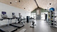 Located at 300 Gatehouse Ln in Odenton, Maryland, this well-appointed fitness center offers a spacious, single-floor layout designed to support a full range of workout routines. The room's most striking architectural feature is its soaring vaulted ceiling clad in light-toned wood paneling, which lends a warm, modern aesthetic while creating an open, airy atmosphere. Recessed and pendant lighting throughout ensures excellent visibility at all hours.

The cardio zone is equipped with multiple treadmills featuring integrated display screens, an elliptical trainer, a stair climber, and stationary cycling bikes, providing diverse options for cardiovascular training. A dedicated strength area houses a multi-station cable machine, a Smith machine with a bench press setup, and a comprehensive rack of dumbbells arranged on a tiered stand, catering to both free-weight and machine-based resistance training.

Large windows and glass-panel doors line the perimeter, flooding the space with natural light and offering views of the surrounding exterior. Full-length mirrors are strategically placed to enhance spatial perception and support proper form during exercise. The floors are finished with durable rubber gym flooring throughout, ensuring safety and comfort. An accent wall in deep green with tasteful artwork adds a refined, contemporary touch to the overall design.