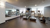Located at 1317 East St in New Britain, Connecticut, this fitness center offers a well-organized and fully equipped exercise facility across a single floor. The space is designed with functionality in mind, featuring durable dark rubber flooring throughout that provides both safety and comfort during workouts. The walls are finished in a neutral light gray tone, complemented by crown molding details and a textured ceiling with recessed lighting that creates a clean, well-illuminated environment. Wall-mounted sconce lighting adds additional brightness throughout the space.

The cardio area is a standout feature, housing multiple treadmills, an elliptical trainer, and a stationary bike, all positioned near windows that allow natural light to filter in. A wall-mounted television is conveniently positioned for viewing during cardio sessions. A large wall mirror spans one side of this area, visually expanding the space and aiding in form checks during exercise.

The strength training section is equally well-appointed, offering a cable machine, a leg press or extension machine, a multi-functional weight rack, adjustable benches, dumbbells, and medicine balls. A smaller mirror is also present in this area to support proper technique. Multiple gray doors provide access to additional rooms or storage areas within the facility, ensuring the space remains organized and accessible.