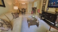 This single-floor residence offers a seamlessly connected living space defined by warm, neutral tones and tasteful décor. The living area serves as the heart of the home, featuring a plush sofa, a glass-top coffee table with decorative candle accents, and a soft area rug that grounds the space with comfort and style. Framed artwork adorns the walls, adding a curated, gallery-like touch to the room. The space flows naturally into a dedicated dining area, illuminated by a chandelier that casts a warm, inviting glow over a classic dining table and chair set. Light-toned tile flooring throughout the shared living and dining areas lends a clean, cohesive aesthetic. Adjacent to the dining area, a breakfast bar with bar-height seating provides an additional casual dining option, connecting seamlessly to the kitchen beyond. The overall ambiance is warm, welcoming, and thoughtfully arranged, making the most of the open layout. The combination of tile and carpeted surfaces, layered lighting, and carefully selected furnishings create an atmosphere that is both functional and aesthetically refined — ideal for everyday living and entertaining alike.