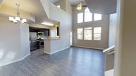 This two-floor space makes a striking first impression with its dramatic vaulted ceilings that soar to impressive heights in the main living area, creating an expansive and airy atmosphere. The open-concept layout seamlessly connects the living room and kitchen, promoting a sense of flow and connectivity throughout the main level. Large windows along the living room wall invite an abundance of natural light and offer pleasant views of the surrounding greenery, while an arched transom window near the ceiling peak adds an elegant architectural touch. A ceiling fan with a light fixture is centrally positioned, providing both comfort and function. The kitchen features dark cabinetry paired with light countertops, stainless steel appliances including a refrigerator, microwave, and gas range, and a convenient pass-through peninsula that doubles as a breakfast bar. The space is finished with smooth grey hardwood-style flooring throughout the main level, complementing the neutral wall tones for a clean, modern aesthetic. The two-story design, combined with the open sightlines from the upper level down to the main floor, adds architectural depth and visual interest to the overall layout.
