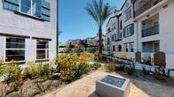 This single-floor residence is situated within a Mediterranean-inspired community characterized by white stucco exteriors, arched architectural details, and teal-shuttered windows. The shared outdoor grounds are thoughtfully landscaped with drought-tolerant plants, flowering shrubs, and a tall palm tree, alongside a pergola and a modern square fire pit feature, creating an inviting communal environment.

Inside, the bedroom serves as a comfortable and well-designed retreat. It features plush wall-to-wall carpeting, neutral-toned walls, and a ceiling fan overhead. A tufted, upholstered headboard anchors the room, complemented by warm bedside lighting. A large window fitted with horizontal blinds allows natural light to filter in while offering views of the surrounding community, including glimpses of a pool area below.

The bedroom connects to a hallway via a wide doorway, along which a built-in dark wood credenza with a large framed mirror adds a refined, functional touch. The room also includes a dedicated closet with built-in shelving, providing organized storage. The corridor leads to additional areas of the residence, maintaining a clean, cohesive aesthetic throughout with white panel doors and recessed lighting.