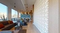 This expansive single-floor amenity space showcases a bold mid-century modern aesthetic, seamlessly blending architectural elegance with vibrant, curated design. Soaring double-height ceilings flood the open-plan interior with natural light through floor-to-ceiling glazing, offering views of the surrounding landscaped exterior. Polished terrazzo floors extend throughout, lending a clean, luminous foundation to the space.

At the heart of the main lounge, dramatic wire-frame pendant light fixtures serve as sculptural focal points overhead, complementing the warm wood ceiling accents. Multiple seating areas are thoughtfully arranged, featuring classic mid-century furnishings including Eames lounge chairs, egg chairs, and plush sofas in earthy and neutral tones, anchored by boldly patterned area rugs.

A long communal bar counter with teal geometric tile detailing and warm wood barstools provides a social gathering point, while a separate lounge room features a striking suspended fireplace, patterned tile flooring, and rich orange upholstered seating beneath a slatted wood ceiling. Decorative breeze-block screens and textured accent walls add depth and visual interest. Glass-walled private offices or meeting rooms are accessible off the main corridor. A built-in bookshelf and stepped platform area with warm wood finishes complete this thoughtfully designed, community-oriented space.