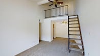 Located in Boulder, this uniquely designed space showcases a striking loft configuration that maximizes vertical space. The main living area is anchored by soaring double-height ceilings, creating an impressive sense of openness and volume. Plush wall-to-wall carpeting in a neutral tone runs throughout, providing warmth and comfort underfoot.

A sleek metal-railed staircase rises along one wall, leading to an elevated mezzanine loft level overhead. This upper level is outfitted with its own ceiling fan and overlooks the main living area through the open metal balustrade railing, making it ideal for use as a sleeping area, home office, or additional lounge space.

On the main floor, a passageway leads to a secondary room, also featuring a ceiling fan, which appears to connect to additional spaces such as a kitchen or hallway. The neutral, light-toned walls throughout enhance the bright and airy atmosphere of the interior.

Ceiling fans on both levels ensure year-round comfort and add a practical, classic touch to the overall aesthetic. The open, multi-level layout offers a dynamic and versatile living environment well-suited to a variety of lifestyle needs.