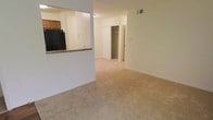 This single-floor space showcases an open-concept layout that connects the living area to the kitchen through a half-wall pass-through counter finished with a dark granite or stone surface. The living area is covered in neutral beige carpet, providing a soft and comfortable underfoot experience throughout the main space. The kitchen, partially visible beyond the pass-through, features dark appliances including a full-size refrigerator and wood-toned cabinetry overhead, offering functional storage solutions.

The walls are finished in a light, neutral tone that contributes to a clean and airy atmosphere throughout the space. A ceiling-mounted HVAC vent is positioned overhead, ensuring efficient climate control. Toward the rear of the living area, a set of white bi-fold closet doors opens to reveal a built-in closet with shelving, providing ample storage. Additional wall outlets are distributed throughout, adding practical convenience.

The overall layout is straightforward and efficient, making good use of the available floor space. The combination of light walls, neutral carpet, and open sightlines creates a bright and functional living environment suitable for a variety of furniture arrangements.