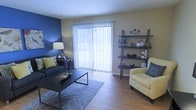 This single-floor space offers a comfortable and thoughtfully designed living environment. The living area serves as the focal point of the home, showcasing warm hardwood-style flooring that adds a sense of continuity and warmth throughout the space. A striking blue accent wall provides a bold design contrast against the neutral beige walls, creating a modern and inviting atmosphere. A large sliding glass door dressed with vertical blinds dominates one wall, allowing an abundance of natural light to pour into the room while offering access to the outdoors. The living area is well-suited for both relaxation and entertaining, with ample room for seating arrangements and decorative furnishings. A lean ladder-style shelving unit positioned near the window provides practical display and storage space. The overall ambiance of the space is bright, clean, and contemporary, with a color palette of blues, yellows, and neutrals that lends a cohesive and welcoming feel. The layout is efficient and functional, making excellent use of the available floor space across the single level.