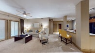 This single-floor residence offers a thoughtfully designed layout with a warm, neutral color palette throughout. The open-concept living area is anchored by plush carpeting, a ceiling fan, and ambient lamp lighting, creating an inviting atmosphere. Bold yellow accent chairs at the kitchen bar counter provide a lively contrast to the soft beige tones. The galley-style kitchen features light wood flooring, warm oak cabinetry, granite countertops, stainless steel appliances including a refrigerator and range, and a built-in dishwasher — offering both style and functionality. A dedicated dining nook showcases a round blue glass table complemented by an elegant gold sunburst mirror, adding a refined decorative touch. The primary bedroom is a serene retreat with carpeted floors, a ceiling fan, natural light from draped windows, and access to a private en suite bathroom complete with a shower/tub combo, dual sinks, and a spacious walk-in closet with built-in shelving. A second bedroom features its own closet and easy access to an additional full bathroom with tile flooring and a shower/tub combination. Throughout the residence, consistent warm-toned walls and neutral finishes create a cohesive and comfortable living environment.