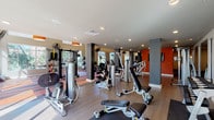 This expansive fitness center is thoughtfully designed to accommodate a full range of workout needs across a single, open floor plan. The space is divided into distinct functional zones, including a dedicated cardio area and a comprehensive strength training section, each outfitted with modern, commercial-grade equipment such as treadmills, stationary bikes, elliptical machines, cable systems, free weights, and resistance machines.

Architecturally, the facility stands out with soaring double-height ceilings in the cardio zone, creating an airy, open atmosphere enhanced by large pendant light fixtures with bold orange shades. Striped, color-blocked carpeting in terracotta and charcoal tones defines the cardio area, while hardwood-style flooring runs throughout the strength training sections, adding warmth and contrast.

Natural light floods the space through generous floor-to-ceiling windows and glass entry doors, offering outdoor views of surrounding greenery. Vibrant orange accent walls and dark structural columns punctuate the interior, lending an energetic and modern aesthetic. Wall-mounted fans ensure comfortable airflow, and strategically placed mirrors amplify the sense of space. Recessed lighting throughout provides consistent, bright illumination. The overall ambiance is motivating, stylish, and highly functional.