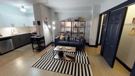 This single-floor space showcases a thoughtfully curated interior with a modern industrial aesthetic. The living area serves as the centerpiece, anchored by a deep navy tufted sofa adorned with an assortment of textured throw pillows. A bold black-and-white striped area rug defines the seating zone, complemented by a sleek metal-and-wood coffee table that adds an industrial yet warm touch. An open metal shelving unit lines the back wall, displaying an eclectic mix of books, decorative objects, and curated accents, lending the space a personalized and lived-in character. A slender floor lamp provides warm ambient lighting that enhances the overall atmosphere. The light hardwood flooring runs throughout, creating a cohesive and airy foundation that contrasts beautifully with the dark furnishings and trim. Crown molding along the ceiling adds a refined architectural detail, bridging the space's contemporary styling with classic design elements. A glimpse into the adjacent kitchen reveals tiled walls and a functional layout that flows naturally from the main living area. The space strikes a balance between sophistication and comfort, making it both visually striking and highly functional.