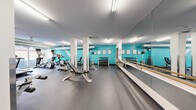 Located at 101 10th St E in Saint Paul, MN, this fitness center offers a thoughtfully designed single-floor workout space that accommodates a wide range of exercise needs. The gym is defined by its bold teal accent walls, crisp white structural columns, and durable gray rubber flooring, creating a clean and motivating atmosphere throughout.

The cardio zone features a row of treadmills positioned along large windows that bring in natural light and provide views of the surrounding streetscape, alongside multiple elliptical trainers and a stationary bike. Exposed silver ductwork along the ceiling adds an industrial character to this section of the gym.

The free weights area is well-stocked with multiple racks of dumbbells and an adjustable weight bench, with workout charts posted directly on the teal wall for easy reference. A full-length mirror spans an entire wall of this section, paired with a barre rail — making the space suitable for stretching, balance training, and barre-style workouts.

Additional strength equipment includes a cable machine, a leg press, and a medicine ball rack with storage for accessories. The space is lit by bright linear fluorescent fixtures, ensuring even, consistent illumination across all workout zones. The fitness center is accessed through a glass-paneled door and is fully self-contained, offering a comprehensive training environment within a compact yet efficiently organized footprint.