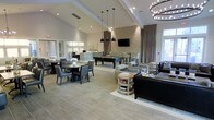 This single-floor amenity space is designed as a versatile social hub, seamlessly blending multiple functional zones within one open-plan layout. The soaring vaulted ceiling creates an airy, expansive atmosphere, complemented by a mix of recessed lighting, sleek pendant lights, and a statement chandelier crafted from a rustic branched frame, adding character and warmth to the modern aesthetic.

The dining area features a round table surrounded by leather-upholstered chairs, ideal for casual gatherings. Nearby, a service counter or kitchenette provides added convenience. Anchoring the center of the space is a full-size billiard table, positioned beneath industrial-style pendant lighting and in front of a textured stone accent wall that houses a wall-mounted flat-screen television. Bar-height seating flanks this area, offering additional casual perches.

A comfortable lounge zone is arranged nearby, featuring plush sofas, accent chairs, and a rustic wooden coffee table atop a neutral area rug. Large windows and glass doors invite natural light throughout. The neutral tile flooring and soft, contemporary color palette of whites, grays, and taupes unify the entire space.
