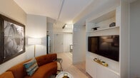 This thoughtfully laid-out single-floor residence offers a cohesive blend of modern finishes and functional design throughout its compact footprint. The living area serves as the heart of the unit, centered around a curved rust-toned sectional sofa, a built-in white media wall with shelving, and track lighting overhead. Light wood-style flooring runs continuously through the space, enhancing the sense of openness.

A dedicated nook adjacent to the living area accommodates a desk and seating area beneath a large window with roller shades, providing natural light and a practical workspace. Framed artwork adorns the soft blue-grey walls, adding a curated aesthetic.

The kitchen is efficiently designed with grey cabinetry, white countertops, and a full suite of stainless steel appliances, including an electric range, over-range microwave, and dishwasher. A stacked washer and dryer unit provides the convenience of in-unit laundry.

The bedroom features a sliding closet door, built-in shelving with an integrated television, and a window with roller shades offering natural light. The bathroom includes a combined tub and shower with a curtain, a vanity with ample counter space, and neutral tiling throughout.