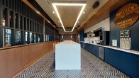 This single-floor amenity space is thoughtfully designed to balance comfort, functionality, and contemporary style. Floor-to-ceiling glass walls along one side of the main lounge flood the interior with abundant natural light and offer expansive outdoor views, creating a seamless connection between inside and outside. The lounge area features polished concrete flooring, soaring exposed ceilings with industrial-style black trusses, and structural columns wrapped in warm wood tones. Seating arrangements include leather sofas, accent chairs, and layered rugs that add color and warmth to the open layout.

A dedicated kitchen area is finished with bold navy cabinetry accented by gold hardware, white quartz countertops, a glossy subway tile backsplash, open wood shelving, and a striking geometric-patterned tile floor. A large central island anchored beneath a sculptural geometric LED light fixture provides additional workspace and gathering space.

Adjacent to the kitchen, a bar area features matching navy cabinetry, a stone-textured accent wall, a large mounted television, and a wood-slat ceiling that adds texture and warmth. The game area includes a shuffleboard table and rustic wall décor, lending a relaxed, recreational character. Green curtains and slatted wood partitions help define zones within the expansive open floor plan, contributing to the cohesive yet versatile design.