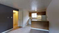 This single-floor residential space offers a practical and functional layout designed for comfortable everyday living. The open-concept design seamlessly connects the kitchen and living area, creating an inviting and efficient use of space. The kitchen is outfitted with warm medium-toned wood cabinetry that provides ample storage, a full suite of appliances including a refrigerator, electric range, and dishwasher, and a countertop peninsula that serves as a natural divider between the kitchen and the adjoining living area. A window above the sink allows natural light to filter into the kitchen, brightening the space. The flooring transitions from easy-to-clean tile in the kitchen area to soft carpeting in the living space, adding warmth underfoot. A textured ceiling runs throughout, lending a classic residential character to the interior. A doorway visible from the main living area leads to additional rooms beyond, suggesting a well-organized floor plan with clear separation between living and private spaces. The neutral wall tones and understated finishes throughout create a clean, versatile backdrop well-suited for a variety of décor styles. Overall, the space presents a straightforward, move-in-ready environment with all essential amenities in place.