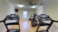 This single-floor fitness space is designed to support a focused cardio workout experience. The room features light-toned hardwood-style flooring that contributes to a clean, energetic atmosphere. Multiple treadmills are positioned toward the front of the room, complemented by an elliptical trainer and a stationary bike, providing a well-rounded selection of cardio equipment. A wall-mounted flat-screen television is positioned for easy viewing during workouts, adding an entertainment element to exercise sessions. Overhead, a ceiling fan with a light fixture helps maintain airflow and comfort throughout the room. Flush-mounted lighting panels ensure the space is well-lit. Crown molding along the upper walls adds a subtle finishing detail. A window with horizontal blinds along the far wall allows for natural light while maintaining privacy. The neutral, light green-toned walls keep the environment calm and uncluttered. Overall, the space is efficiently arranged to maximize usable floor area while providing a comfortable, practical setting for regular cardiovascular training.