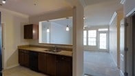 This single-floor residence showcases a thoughtfully designed open-concept layout that seamlessly connects the kitchen and living areas. The kitchen is equipped with rich dark wood cabinetry, granite countertops, and a double-basin sink, all anchored by a partial wall with a pass-through opening that enhances the sense of openness. Two sleek pendant lights hang above the counter, adding focused task lighting while contributing to the modern aesthetic. Black appliances complement the dark cabinetry for a cohesive look. Crown molding details throughout the space lend a polished, refined character. The adjoining living area features neutral-toned carpeting and benefits from generous natural light streaming through multiple large windows along the far wall, creating a bright and welcoming atmosphere. A ceiling fan is installed in the living area, adding both function and comfort. The overall design balances practicality with style, featuring clean lines, neutral tones, and quality finishes that create a cohesive and inviting environment throughout the space.