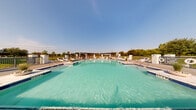 Located at 6000 Eldorado Pkwy in Frisco, Texas, this outdoor amenity area offers a resort-inspired experience designed for relaxation and recreation. The centerpiece is a generously sized swimming pool with crystal-clear turquoise water, framed by a wide surrounding deck finished in elegant paver stonework. Multiple lounge chairs are arranged throughout the deck, providing ample space for sunbathing under the open Texas sky.

At the rear of the pool area, a striking pergola structure with striped drapery panels serves as a shaded retreat, adding architectural character while offering a comfortable respite from the sun. Stone pillar accents and decorative planters punctuate the perimeter, lending the space a polished, upscale aesthetic.

Adjacent to the main pool is a dedicated spa and shallow wading area, complete with water features that create a soothing atmosphere. A well-appointed outdoor patio extends from the clubhouse building, which features arched entryways, white stone detailing, and large windows. The patio includes seating and dining areas ideal for casual gatherings.

The surrounding landscape is thoughtfully maintained, with manicured greenery, trees, and open lawn areas visible beyond the fenced perimeter. The entire space is bathed in natural light and open to panoramic views of the sky, making it a vibrant and inviting outdoor destination.