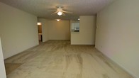 This single-floor space features an open-concept layout centered around a spacious living area finished with wall-to-wall carpeting in a neutral beige tone. A ceiling fan with an integrated light fixture is mounted to the textured popcorn ceiling, providing both air circulation and ambient lighting throughout the main living space.

A pass-through window connects the living area to the kitchen, which is visible from the main room and features white cabinetry and appliances, including a microwave. This design element enhances the sense of openness while maintaining a functional separation between the cooking and living areas.

A hallway on the left side of the space leads to additional rooms, with a glimpse of what appears to be a bathroom or secondary space visible from the main area. The walls are finished in a light neutral tone, complementing the carpeted floors and contributing to a cohesive, understated aesthetic throughout.

Overall, the space presents a simple, functional layout with an emphasis on open living, offering a practical and adaptable interior suitable for a variety of furnishing arrangements.