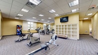This fitness center offers a well-equipped, single-floor layout designed to accommodate a wide range of workout needs. The space features a drop ceiling with recessed fluorescent lighting panels that provide consistent, even illumination throughout. Warm yellow walls contribute to an energizing atmosphere, while multiple windows along the perimeter invite natural light and offer views of the surrounding outdoor greenery.

The cardio section includes a treadmill, elliptical trainers, stationary bikes, and a rowing machine, each positioned on protective rubber mats. A dedicated strength training area houses a multi-station cable machine and weight benches, complemented by a rack of free weights and resistance equipment. Full-length mirrors are strategically placed to support proper form during exercise.

A wall-mounted flat-screen television is prominently positioned for viewing during workouts. Built-in shelving units provide convenient storage for personal belongings and fitness accessories. A beverage vending machine near the entrance adds a practical amenity. The carpeted flooring throughout the main area ensures comfort underfoot, while a separate section features hard flooring suited for stretching and floor exercises. The layout is open and thoughtfully organized to maximize usability.