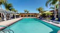 Located at 17301 Keelson Ln in Huntington Beach, California, this outdoor amenity area embodies the relaxed, sun-soaked lifestyle of Southern California. At the heart of the space is a generously sized swimming pool with clear, blue-green water, framed by brick-accented coping and accessible via a stainless steel entry rail. The pool area is enclosed by a black wrought-iron security fence, providing both safety and a clean aesthetic boundary.

Surrounding the pool is a spacious concrete deck furnished with blue lounge chairs and vibrant green market umbrellas, offering ample space for sunbathing and relaxation. A standout feature is the white pergola structure positioned along one side of the pool deck, fitted with flowing sheer curtains and stylish navy blue outdoor seating sets — creating a shaded cabana retreat ideal for socializing or unwinding out of direct sun.

Towering palm trees are planted throughout the area, adding a tropical ambiance and natural privacy screening. The adjacent single-story building, finished in warm earth tones, houses what appears to be a pool support facility with direct access to the outdoor area. The overall design is cohesive and well-maintained, combining functional amenities with an inviting, resort-inspired atmosphere perfectly suited to the Southern California climate.