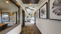 This thoughtfully designed business center combines functionality with contemporary style across a single floor. An art-lined corridor with patterned carpeting and large framed mirrors serves as an inviting entry, transitioning seamlessly into the various areas of the space.

The central lounge area is a standout, featuring soaring vaulted ceilings accented by exposed wooden beams and a striking sputnik chandelier. A generous sectional sofa in soft gray and a round wooden coffee table anchor the space, while lime-green accent chairs positioned near large multi-pane windows create cozy nooks flooded with natural light and views of the surrounding landscape.

Adjacent to the lounge, a dining area offers a round table with modern chairs, surrounded by additional windows and curated wall art, making it well-suited for informal meetings or collaborative work sessions.

The dedicated workstation area features built-in desks, ergonomic seating, and ample natural light streaming through large windows overlooking greenery. A printer station and storage cabinet complete the practical amenities.

Throughout the space, an extensive gallery-style art collection adorns the walls, adding visual depth and personality. The consistent use of patterned area rugs, neutral tones, and green accents ties the entire business center together in a cohesive, professional yet welcoming aesthetic.