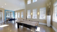 This game room is designed for both leisure and social gatherings, immediately drawing attention with its dramatically high ceilings that lend an open, airy quality to the space. A full-size billiard table serves as the centerpiece, positioned to take full advantage of the generous floor space, with cue storage conveniently nearby.

The room is bathed in natural light from multiple large windows arranged along the walls, including a row of clerestory windows near the ceiling that amplify the sense of height and brightness. Roller shades on each window allow for flexible light control. Wall-mounted sconce lighting complements the natural illumination, adding a warm ambiance during evening hours.

The walls are finished in a neutral warm beige, contrasted by a vibrant teal accent wall visible in the adjoining area, which adds a contemporary pop of color. Polished concrete floors throughout are both durable and visually clean, well-suited to a high-traffic recreational space. A seating area with dark chairs is positioned near the windows, offering a relaxed spot to gather. Overall, the space balances functionality with a modern, welcoming design aesthetic.