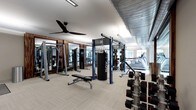 This fitness center offers a thoughtfully designed single-floor layout that accommodates a full range of training activities. The space features high white ceilings accented by exposed industrial-style ductwork, giving the room a modern yet functional character. A large ceiling fan ensures comfortable air circulation throughout the workout area.

The facility is outfitted with an impressive selection of equipment, including a multi-station cable and weight machine, a heavy punching bag suspended from a freestanding blue power rack, a dumbbell rack with multiple weight selections, and a flat-to-incline adjustable bench. Cardio equipment, including treadmills, is positioned along the perimeter to maximize open floor space.

Floor-to-ceiling mirrors line one wall, enhancing the sense of openness and allowing users to monitor their form during exercise. The flooring consists of light-toned, durable tile-style material well-suited for high-traffic fitness use. Natural light filters in through windows at one end of the room, brightening the space and creating an energizing atmosphere. The combination of thoughtful equipment placement, ample space between stations, and a clean, contemporary design makes this fitness center a well-rounded facility for a variety of fitness routines.