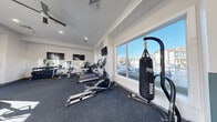 Located at 901 W Little Creek Rd in Norfolk, Virginia, this fitness center offers a well-equipped, single-floor workout space designed to support a comprehensive exercise routine. The interior features clean white walls, dark carpeted flooring, and a bright, open layout bathed in natural light streaming through large sliding windows that overlook the surrounding community and outdoor pool area.

The cardio zone is well-stocked with multiple treadmills, elliptical machines, and a recumbent stationary bike, providing ample options for aerobic training. A freestanding heavy bag and MMA-style punching bag station add a combat fitness element to the space. For strength training, a dedicated free weights area includes a full dumbbell rack with a wall-mounted mirror, along with weight benches and rowing machines.

Multiple wall-mounted flat-screen televisions are positioned throughout the room, allowing members to stay entertained during workouts. Ceiling fans and recessed lighting contribute to a comfortable and well-lit environment. Two entry points — including a glass door with exterior access — ensure easy flow in and out of the space. A hand sanitizer station and posted fitness center rules reflect a commitment to cleanliness and community standards. Overall, this fitness center delivers a functional, modern workout environment.