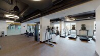 This fitness center offers a comprehensive workout environment designed to accommodate a wide range of exercise needs. The space features an industrial-modern aesthetic, with exposed ductwork and structural elements set against a dark ceiling, complemented by bold circular pendant lighting that illuminates the floor below.

The cardio area is generously outfitted with a row of treadmills, elliptical trainers, and stationary bikes, all positioned along large windows that invite ample natural light and outdoor views. Adjacent to this section, a dedicated strength training zone houses a variety of cable machines, a leg press, weight benches, and a full dumbbell rack, all reflected in floor-to-ceiling mirrors that enhance the sense of space.

A separate studio area features warm hardwood flooring and a striking graphic mural wall in bold yellow, black, and white tones, with full-length mirrors and a mounted flat-screen display — ideal for group classes or individual stretching and training sessions. Towel service is conveniently available near the free weights section, adding a refined amenity touch.

Throughout the fitness center, the combination of neutral carpeted flooring in the equipment zones, white structural columns, and accent lighting creates a clean, motivating atmosphere well-suited for daily use.