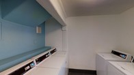 This single-floor laundry facility is a practical and functional space designed to accommodate multiple users efficiently. The room features a row of commercial-grade top-load washing machines and dryers neatly arranged along one wall, providing ample capacity for regular laundry needs. Each machine is equipped with digital control panels, reflecting modern, user-friendly functionality.

The interior design is simple yet clean, with white walls on one side contrasting against a bold teal accent wall that adds a touch of color and visual interest to the otherwise utilitarian space. The ceiling is textured and painted white, contributing to the bright, open feel of the room. Dark tile flooring runs throughout, offering a durable and easy-to-maintain surface well-suited to a high-traffic laundry environment.

A recessed structural element near the teal wall creates a defined nook that houses the primary row of machines, while additional units are positioned along the adjacent white wall, maximizing the use of available space. The overall layout is straightforward and efficient, prioritizing accessibility and ease of use. The space presents a clean, well-maintained environment suitable for shared residential or commercial laundry use.