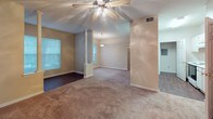 This single-floor residence offers a thoughtfully arranged layout that balances open communal areas with private retreats. The living and dining areas are connected by an open flow, distinguished by decorative white columns that add a subtle architectural detail. The living room features plush carpeting, a ceiling fan with integrated lighting, and windows that invite natural light and outdoor views. A pendant light fixture marks the dining area, which also benefits from generous window coverage. A glass-paneled exterior door provides additional natural light and outdoor access.

The galley-style kitchen is efficiently organized, featuring white upper and lower cabinetry, white countertops, a full-size refrigerator, an electric range, and dark hardwood-style flooring that contrasts cleanly with the bright cabinetry. A doorway at the kitchen's end leads to a utility or storage area with visible shelving and an electrical panel.

The bedroom is carpeted and equipped with a ceiling fan, offering a comfortable and quiet atmosphere. It is served by a spacious walk-in closet outfitted with multiple wire shelving units and hanging rods for ample storage. The bathroom features dark hardwood-style flooring, a wide vanity with white cabinetry, a large mirror, a lighted fixture, and a window for natural light.