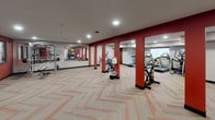 This fitness center is a comprehensively equipped exercise facility designed to support a full range of workout routines. The space is defined by a striking interior palette featuring bold red accent walls and columns contrasted against neutral beige tones, creating an energetic yet focused atmosphere. Patterned herringbone carpet in warm tones with orange detailing covers the floor throughout, adding visual interest underfoot.

The cardio section is outfitted with multiple treadmills and elliptical machines, as well as stationary and cycling bikes, providing ample options for aerobic exercise. A dedicated free weight area features a tiered dumbbell rack stocked with a full range of weights, positioned against the vivid red accent wall. Additional strength and functional training equipment is distributed throughout the open floor plan.

A mirrored wall section expands the visual depth of the space and supports stretching and form-checking during workouts. A collection of exercise balls in various sizes is neatly organized in one corner, rounding out the functional training offerings. Recessed lighting illuminates the entire area evenly, and structural columns with red detailing reinforce the room's bold design identity. The layout is open and spacious, allowing for comfortable movement between equipment stations.