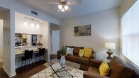 This 2-bedroom, 2-bathroom single-floor residence is thoughtfully designed to maximize comfort and functionality. The open-concept layout seamlessly connects the living area to the kitchen, creating an inviting and cohesive space. The living room features warm hardwood-style flooring, neutral-toned walls, and a ceiling fan with integrated lighting, contributing to a bright and airy atmosphere. A glass coffee table and plush area rug anchor the seating area, which is bathed in warm light from both overhead and accent fixtures. The kitchen is equipped with stainless steel appliances, including a full-size refrigerator and microwave, and features a breakfast bar with seating — ideal for casual dining or entertaining. Pendant lighting above the bar adds a modern touch. A hallway visible from the main living area leads to the bedrooms and additional living spaces, maintaining a clear and practical flow throughout the home. The overall ambiance is warm and welcoming, with a neutral color palette and modern finishes that provide a versatile backdrop suited to a variety of interior styles.