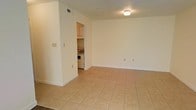 This single-floor space offers a straightforward and functional layout designed for comfortable everyday living. The main living area is finished with large, light-toned ceramic tile flooring that extends throughout, creating a bright and cohesive feel underfoot. The walls are painted in a clean, neutral white, contributing to an open and airy atmosphere. A ceiling-mounted light fixture provides consistent overhead illumination throughout the space.

A partial wall partition separates the living area from the kitchen, which is accessible through an open doorway. The kitchen is compact yet practical, featuring a freestanding gas range and an in-unit washer and dryer, offering added convenience. Overhead cabinetry in the kitchen provides essential storage space.

Electrical outlets are positioned along the walls, and a ceiling vent ensures proper air circulation throughout the unit. The overall design is simple and utilitarian, with clean lines and minimal ornamentation, making it easy to personalize and furnish. This space is well-suited for those seeking a low-maintenance, move-in-ready environment with essential amenities efficiently arranged within a single level.