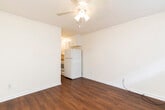 This single-floor unit offers a straightforward, functional layout finished consistently throughout with rich, dark hardwood-style flooring and crisp white walls trimmed with simple crown molding and baseboards. The main living area is open and simply appointed, featuring a ceiling fan with light fixture overhead, a window with a wall-mounted AC unit, and a standard entry door providing natural light and direct access. Crown molding adds a subtle architectural detail throughout the space.

The kitchen is accessed directly from the living area and is equipped with white cabinetry, a white refrigerator, and a white range, creating a clean, cohesive look. Under-cabinet lighting brightens the workspace, and the compact layout makes efficient use of the available area.

Adjacent to the living space, a bedroom receives natural light through a window overlooking an outdoor fence line, and is fitted with a ceiling fan and light combination, keeping the room comfortable and well-lit.

The bathroom is cleanly finished in white throughout, featuring a tub-shower combination with built-in shelving, a toilet, and continued hardwood-style flooring. A ceiling vent provides ventilation. Overall, the unit presents a clean, no-frills interior with consistent finishes across all rooms.