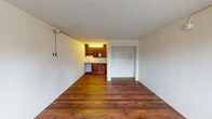 This single-floor unit offers a straightforward, functional layout highlighted by rich, dark hardwood flooring that runs throughout the main living area, creating a warm and cohesive feel underfoot. The open living space is bright and clean, with white walls and a textured ceiling that add a classic character to the interior. A wall-mounted conduit runs along the upper perimeter, and a wall unit provides climate control near the front entry door, which is accompanied by a window that allows natural light to filter in.

The kitchenette is tucked neatly into one corner of the unit, featuring warm medium-toned wood cabinetry both above and below the counter, open shelving for storage, a single-basin sink, and space for a compact appliance such as a dishwasher or refrigerator. Adjacent to the kitchen area is a dedicated closet nook fitted with a hanging rod, offering practical everyday storage.

The bathroom is tiled throughout and includes a wall-mounted sink with a wood-framed mirror cabinet above, a toilet, and a full soaking tub with an overhead shower. A distinctive teal and navy checkerboard tile border accents the bathtub surround, adding a pop of color and a retro charm to the otherwise neutral space. Overall, the unit presents a clean, efficient living environment with functional amenities throughout.