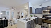 This single-floor residence features a thoughtfully designed open-concept layout with a seamless flow between the kitchen, living, and bedroom areas. Light-toned hardwood-style flooring runs throughout, lending a clean and cohesive aesthetic across all spaces.

The kitchen is a standout feature, equipped with slate-gray shaker-style cabinetry, quartz countertops with a subtle veining pattern, and stainless steel appliances including a range, over-the-range microwave, refrigerator, and dishwasher. A peninsula with a double stainless steel sink and bar seating provides both functional workspace and a casual dining option. A white tile backsplash completes the modern look.

The living area is bright and open, benefiting from natural light through multiple windows fitted with horizontal blinds. A ceiling fan is centrally positioned overhead. The space connects directly to the bedroom corridor, where a decorative entry adds visual interest.

The bedroom is generously sized with multiple windows allowing ample natural light, crown molding details, and a ceiling fan. The en-suite bathroom features gray cabinetry with quartz countertops, recessed lighting, and a large mirror. Adjacent to the bathroom, a hallway with closet access enhances storage and privacy within the unit.