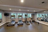Located at 414 Point Return Dr in Ballwin, MO, this fitness center offers a thoughtfully organized exercise space designed to support a wide range of workout routines. The single-level layout is divided into distinct functional zones, maximizing utility throughout the room.

The cardio area is lined with multiple treadmills, an elliptical trainer, a stationary cycle, and a rowing machine, all positioned along a wall of windows that frame lush, green outdoor views. Large mirrors along one wall visually expand the space and allow users to monitor their form during exercise.

The strength training section features a multi-functional cable machine, a squat/power rack with weight plates, a dumbbell rack with an assortment of free weights, kettlebells, a weight bench, and a foam roller for recovery. A wall-mounted exercise mat provides an additional option for floor-based training.

Completing the space is a dedicated flexibility and functional fitness area equipped with a stability ball rack holding multiple sized balls and a selection of medicine balls. Recessed lighting throughout provides consistent, even illumination, while the durable speckled rubber flooring ensures safety and easy maintenance. A bulletin board and sanitation station near the entrance reflect thoughtful amenities for users of this well-appointed community fitness facility.