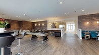 This lounge is a well-appointed social space designed to balance comfort and entertainment. The centerpiece of the room is a substantial billiards table finished in dark felt, anchored by a sturdy trestle-style base that reinforces the space's sophisticated aesthetic. Wide-plank hardwood flooring in a weathered gray tone runs throughout, complementing the warm, reclaimed wood accent wall that adds texture and depth to one side of the room.

Seating is thoughtfully arranged to encourage conversation and relaxation, with a tufted leather sectional sofa and additional lounge chairs positioned nearby. Recessed ceiling lighting provides even illumination across the space, while decorative wall sconces along the wood-paneled wall cast a warmer, ambient glow. A statement chandelier marks the transition toward an adjacent corridor, which leads to a bright entryway framed by large glass doors that invite natural light deep into the interior.

The open layout flows naturally between the recreational area and the passageway beyond, creating a sense of connectivity within the building. The overall design is modern and refined, with a neutral palette and carefully chosen materials that give the lounge a polished, welcoming character.