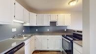 Located at 1750 20th Avenue Dr NE in Hickory, North Carolina, this single-floor residence offers a well-organized layout designed for comfortable everyday living. The kitchen is outfitted with ample white cabinetry providing generous storage, gray countertops, a stainless steel sink, a built-in dishwasher, and a black electric range, all set against a cool blue-gray backsplash and durable wood-look flooring. The open living area is spacious and filled with natural light, featuring the same wood-look flooring and neutral warm-toned walls. Sliding glass doors lead directly to a private balcony that overlooks a wooded outdoor setting, bringing in a pleasant sense of openness. A dedicated bedroom offers plush carpeting and a large window that allows abundant natural light to fill the room. The bathroom is clean and functional, featuring a full tub-shower combination, a white vanity with a marble-style countertop, and wood-look tile flooring. A separate laundry room includes washer and dryer hookups, a utility drain, and houses the electrical panel, adding practical convenience. Textured ceilings are consistent throughout the space, contributing to its classic residential character.