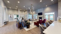 This single-floor communal space is designed with a contemporary aesthetic that balances sophistication and comfort throughout its open, generously scaled layout. The main gathering area commands attention with dramatically high ceilings and a striking chandelier featuring spherical glass globes suspended from a brass frame, casting warm, ambient light across the room. Wood-look flooring flows consistently throughout, anchoring the space in a warm, neutral palette of gray, taupe, and white.

A prominent stone-clad fireplace serves as a focal point along one wall, topped by a wall-mounted television and flanked by white-trimmed glass doors that invite natural light from multiple directions. Multiple seating arrangements are thoughtfully distributed across the main lounge, incorporating gray sofas, jewel-toned burgundy and plum accent chairs, and patterned area rugs.

Stone-faced columns add architectural character and define zones within the open floor plan. A separate lounge nook features a tray ceiling, large wraparound windows with floor-length curtains, and cozy upholstered seating around an octagonal marble-topped coffee table. The kitchen area includes stainless steel appliances, dark wood cabinetry, and a white countertop island, providing practical amenities. Adjacent office or meeting spaces are accessible through French doors, completing the functional layout.
